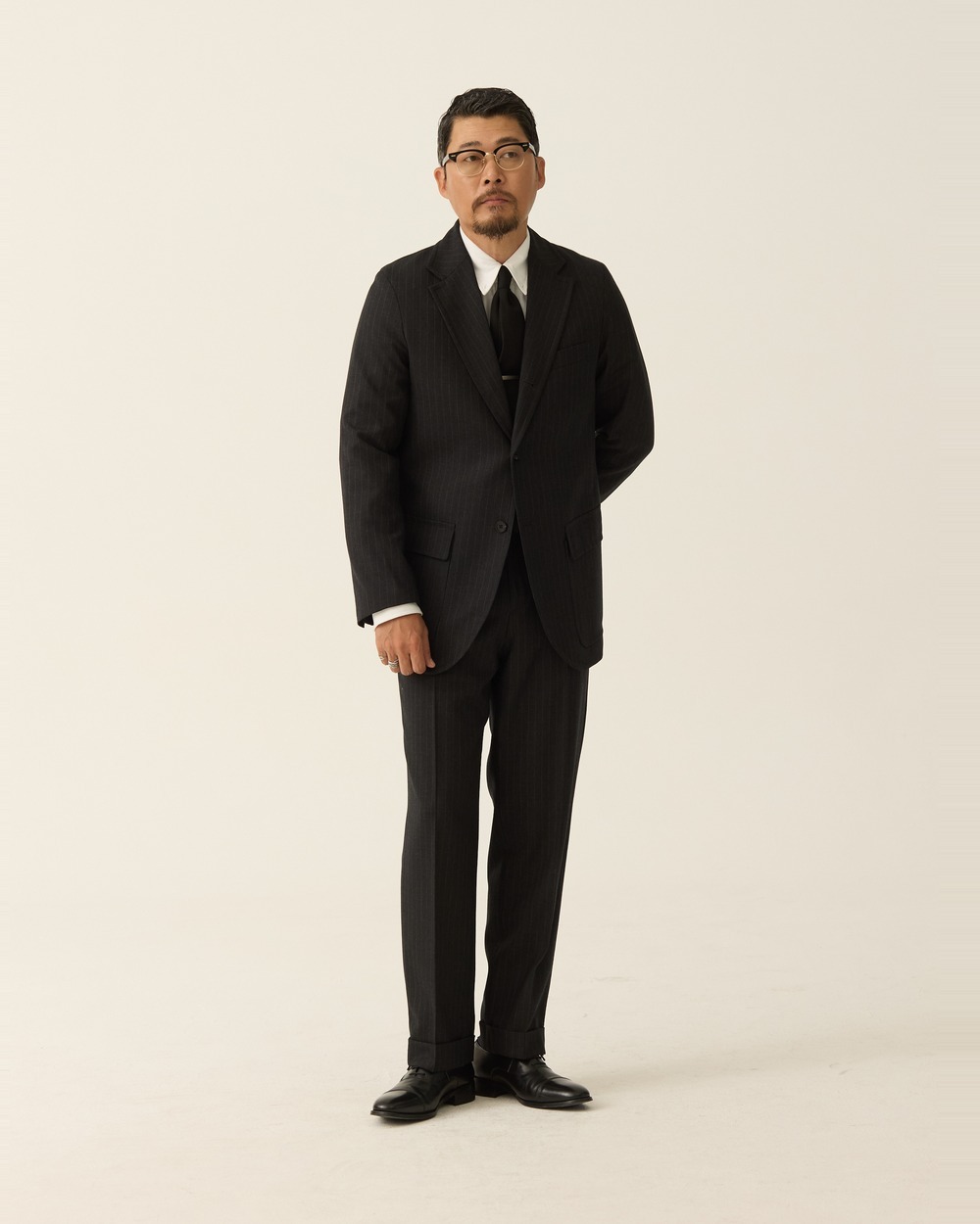 Full-length view of a man in a buttoned charcoal pinstripe sack suit with a white button-down shirt and a dark tie, one hand in his trouser pocket