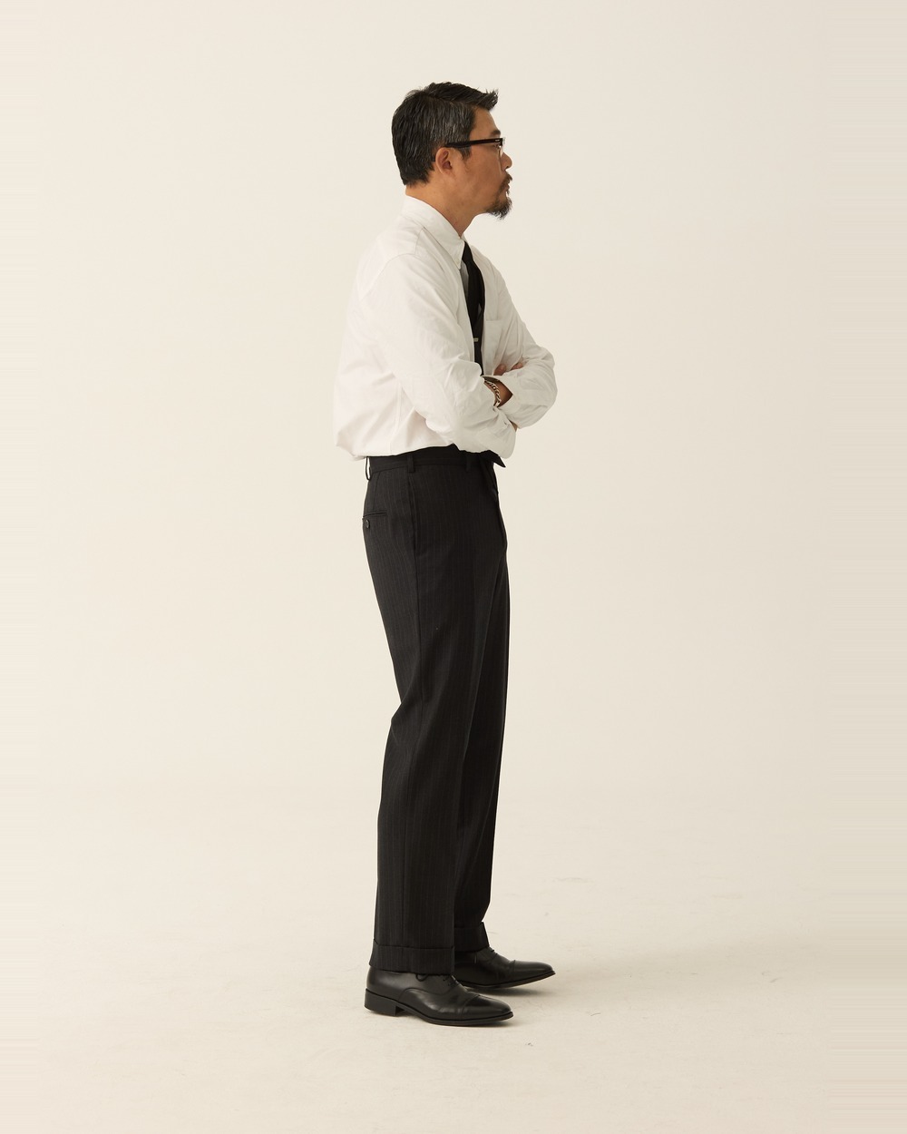Side view of a man in charcoal pinstripe trousers, a tucked-in white shirt and a dark tie, arms folded, with no jacket