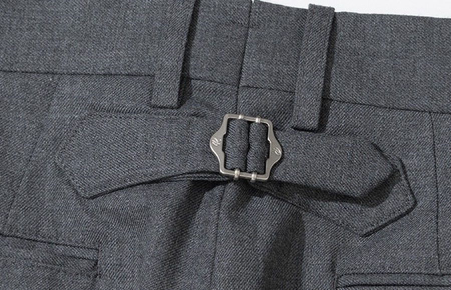 Close-up of the metal cinch-back buckle on the rear waistband of grey wool trousers, with the fabric tab it tightens
