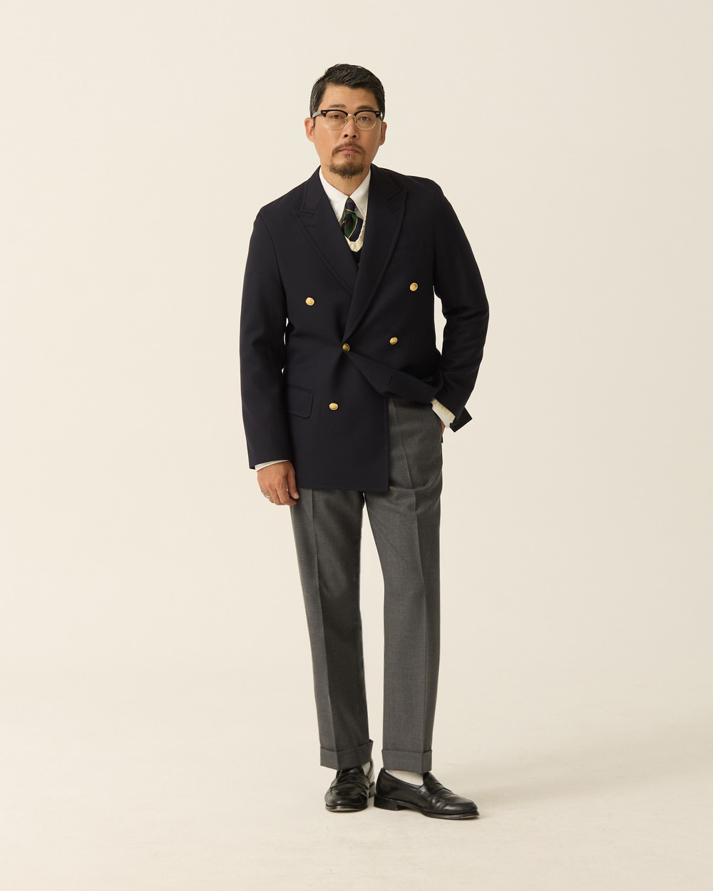 Mid-grey wool trousers with turn-up cuffs worn under a navy double-breasted brass-button blazer with a tartan tie and black loafers