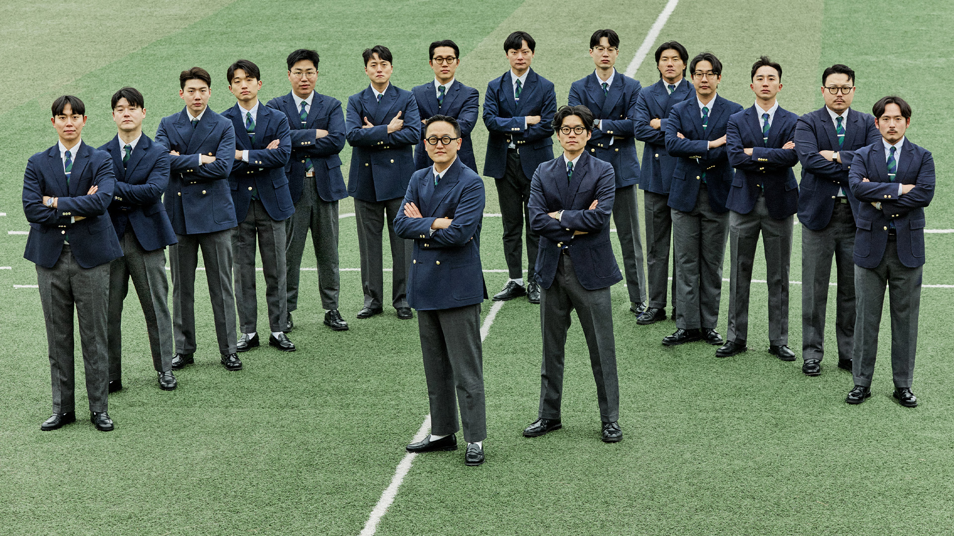 Seventeen men on a football pitch, all in navy blazers with striped ties and mid-grey wool trousers with black shoes