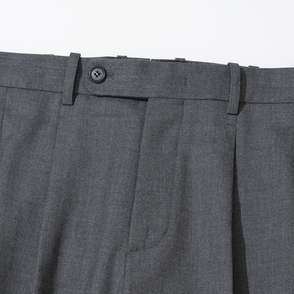 Front waistband detail of grey wool trousers showing a forward-facing pleat, an extended tab closure and a single button
