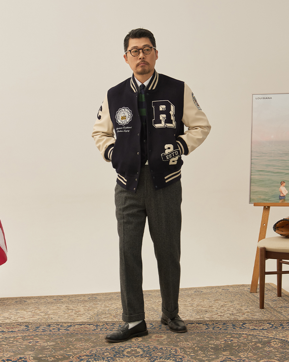 A navy varsity jacket worn over grey wool trousers with turn-up cuffs, a striped tie and black loafers with white socks