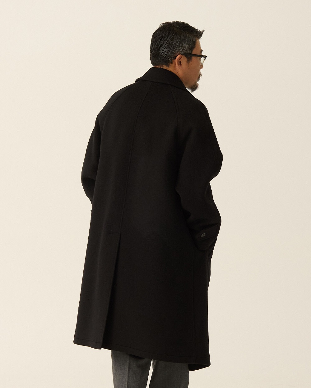 Back view of a man in a black brushed wool balmacaan coat, showing the raglan seams and the vent