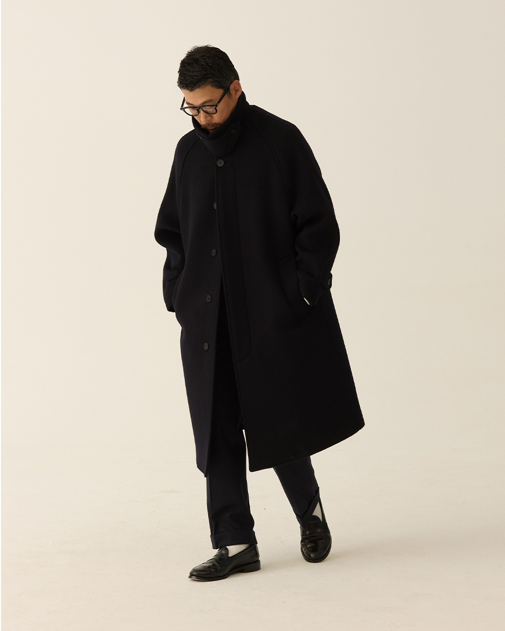 Man in a black brushed wool balmacaan coat worn over a white shirt with blue jeans and black loafers