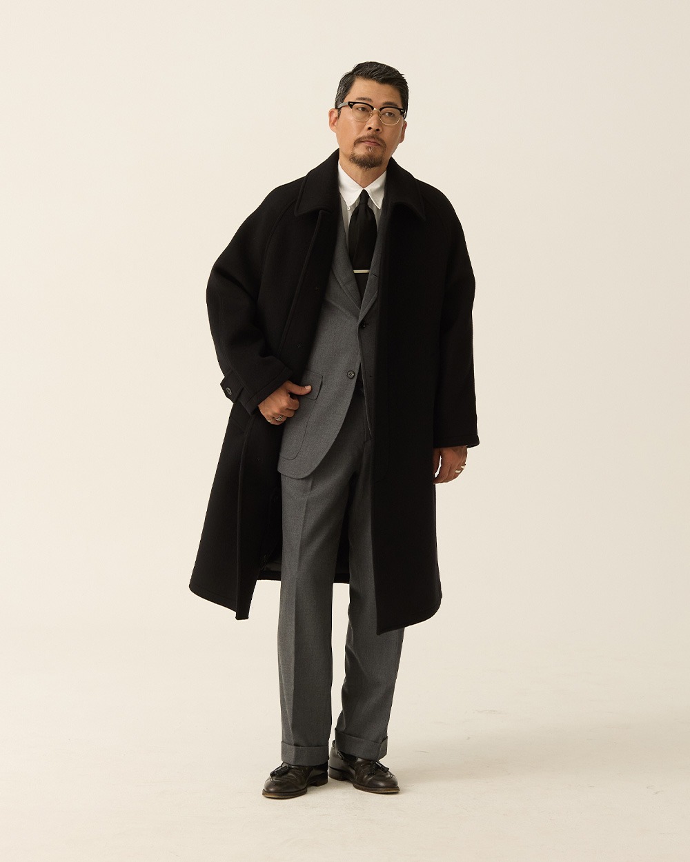 Three-quarter view of a man in a black brushed wool balmacaan coat worn open over a grey suit, white shirt and dark tie