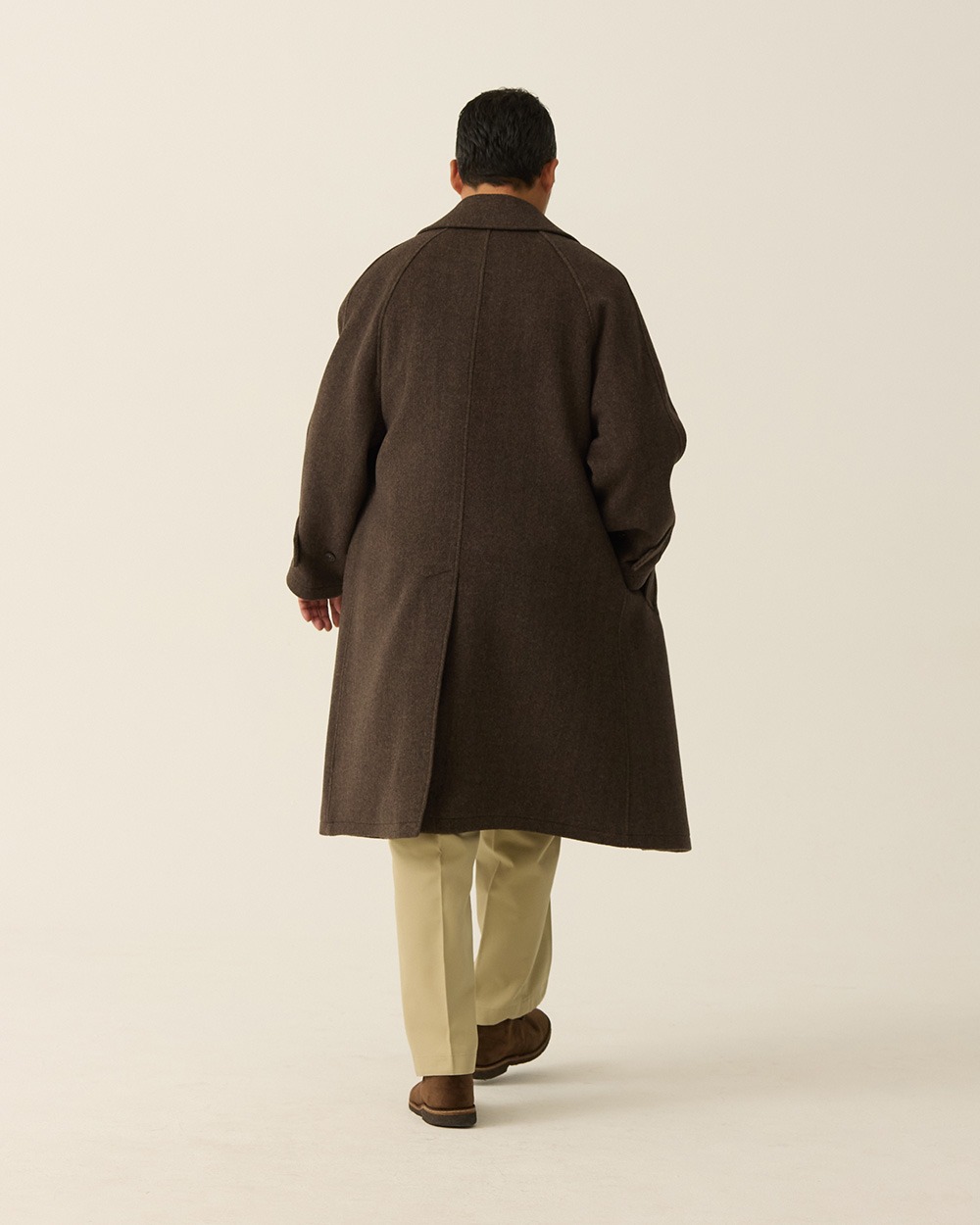 Back view of a man in a brown herringbone balmacaan coat with khaki trousers, showing the raglan seams and vent