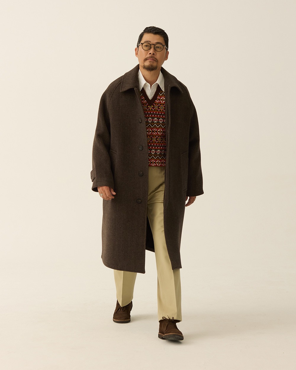 Man wearing a brown herringbone balmacaan coat open over a Fair Isle vest, shirt and tie, with khaki trousers and brown suede shoes