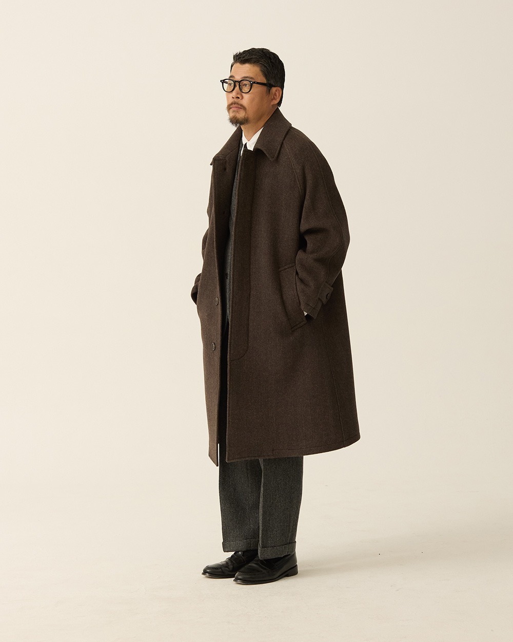 Man in a brown herringbone balmacaan coat worn open over a grey suit, white shirt and tie, with black shoes