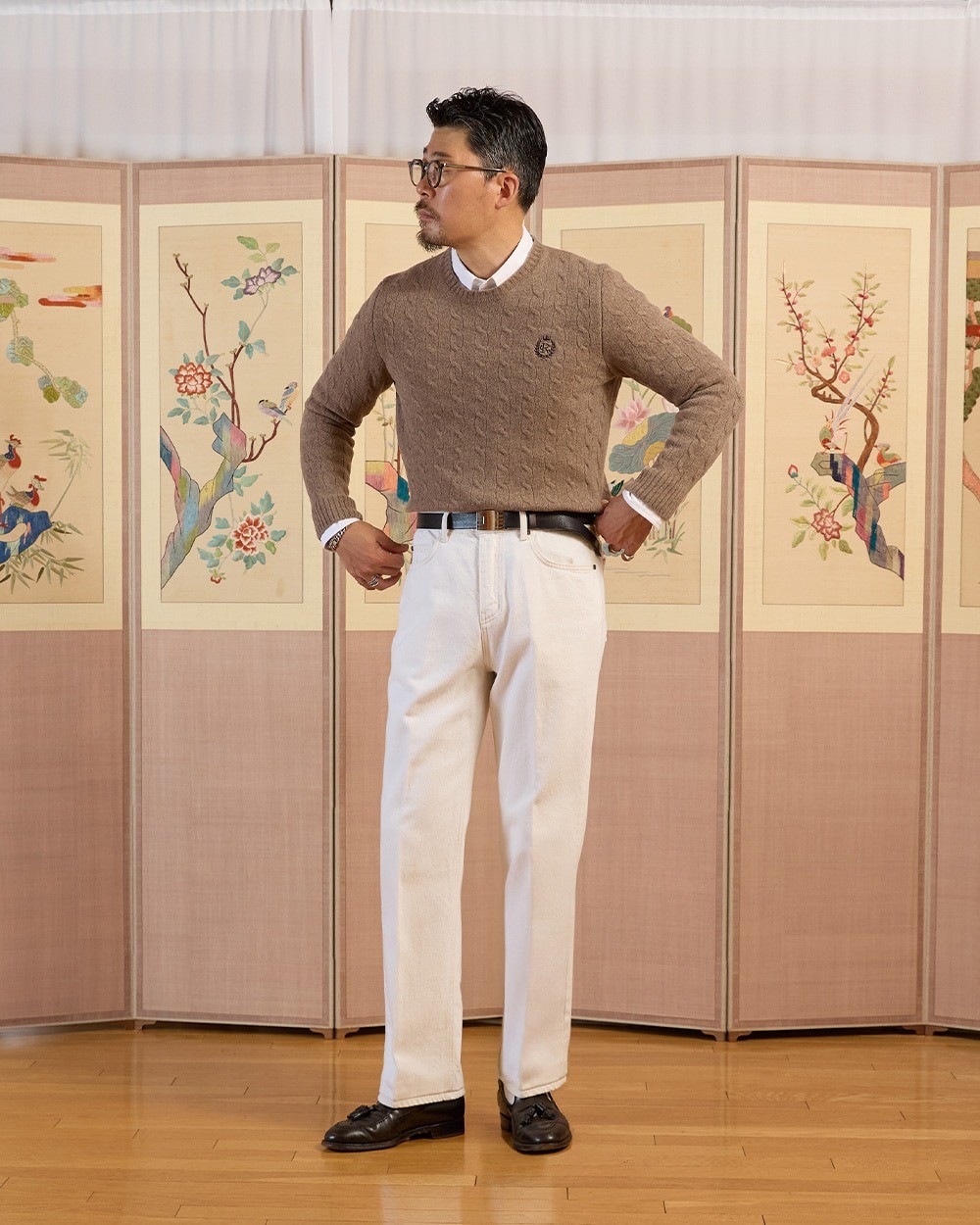 Man in a beige cable knit crewneck sweater over a white button-down collar, cream jeans with a dark leather belt and black tassel loafers, standing in front of a painted folding screen
