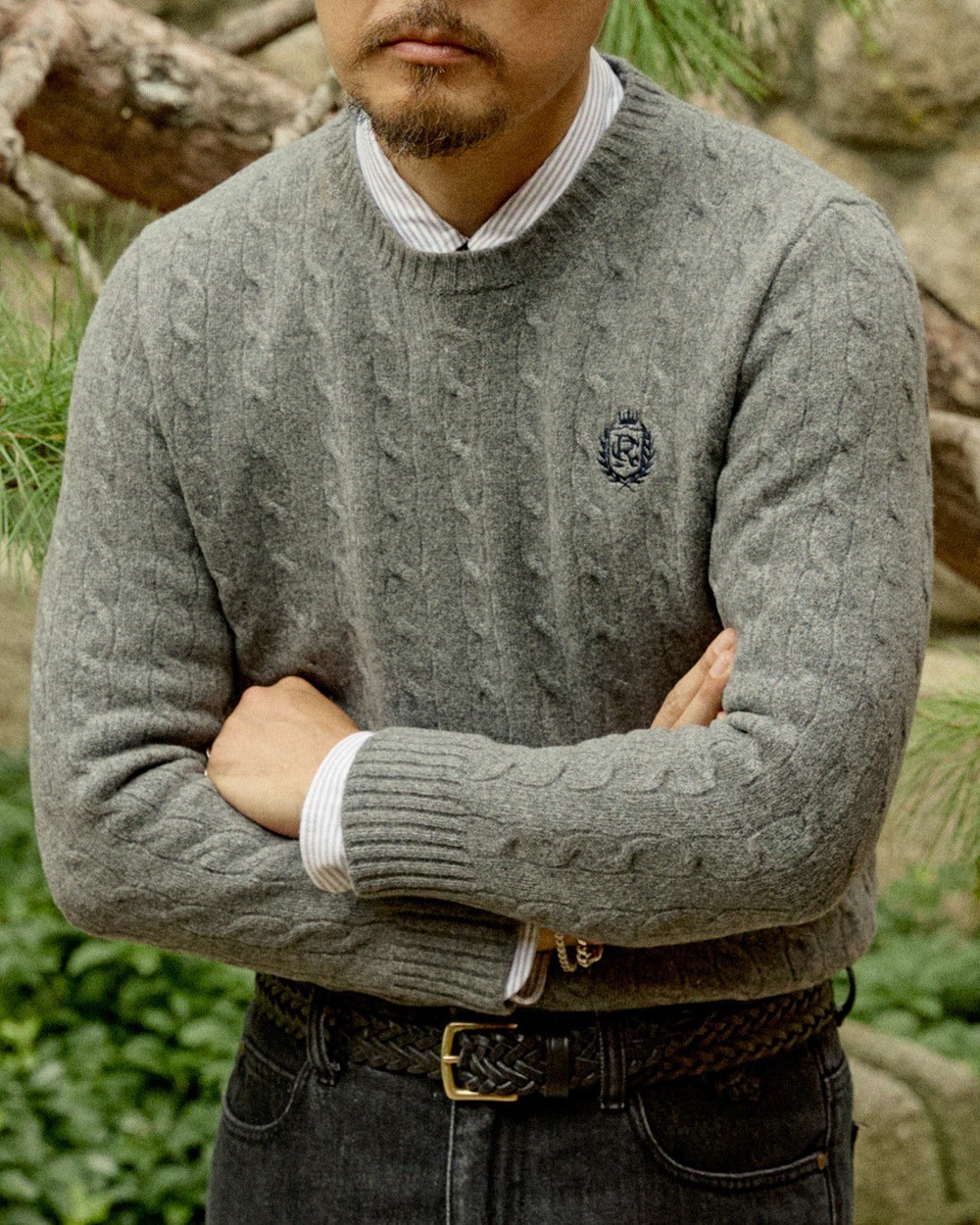 Man with arms crossed in a grey cable knit crewneck sweater over a striped button-down shirt, the ribbed hem stopping at a braided leather belt over dark jeans