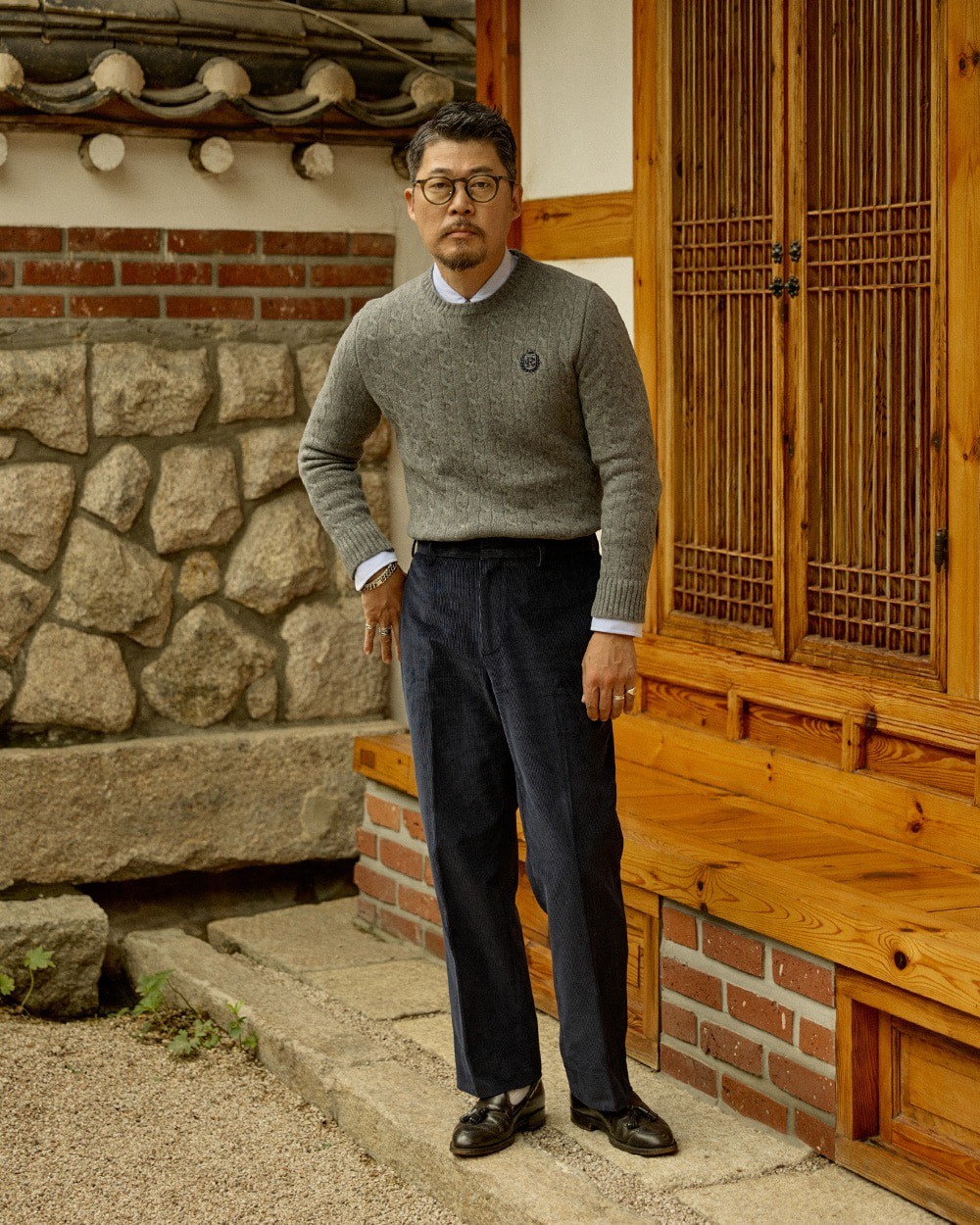 Man in a grey cable knit crewneck sweater over a white collar, with navy corduroy trousers and dark tassel loafers, in a hanok courtyard