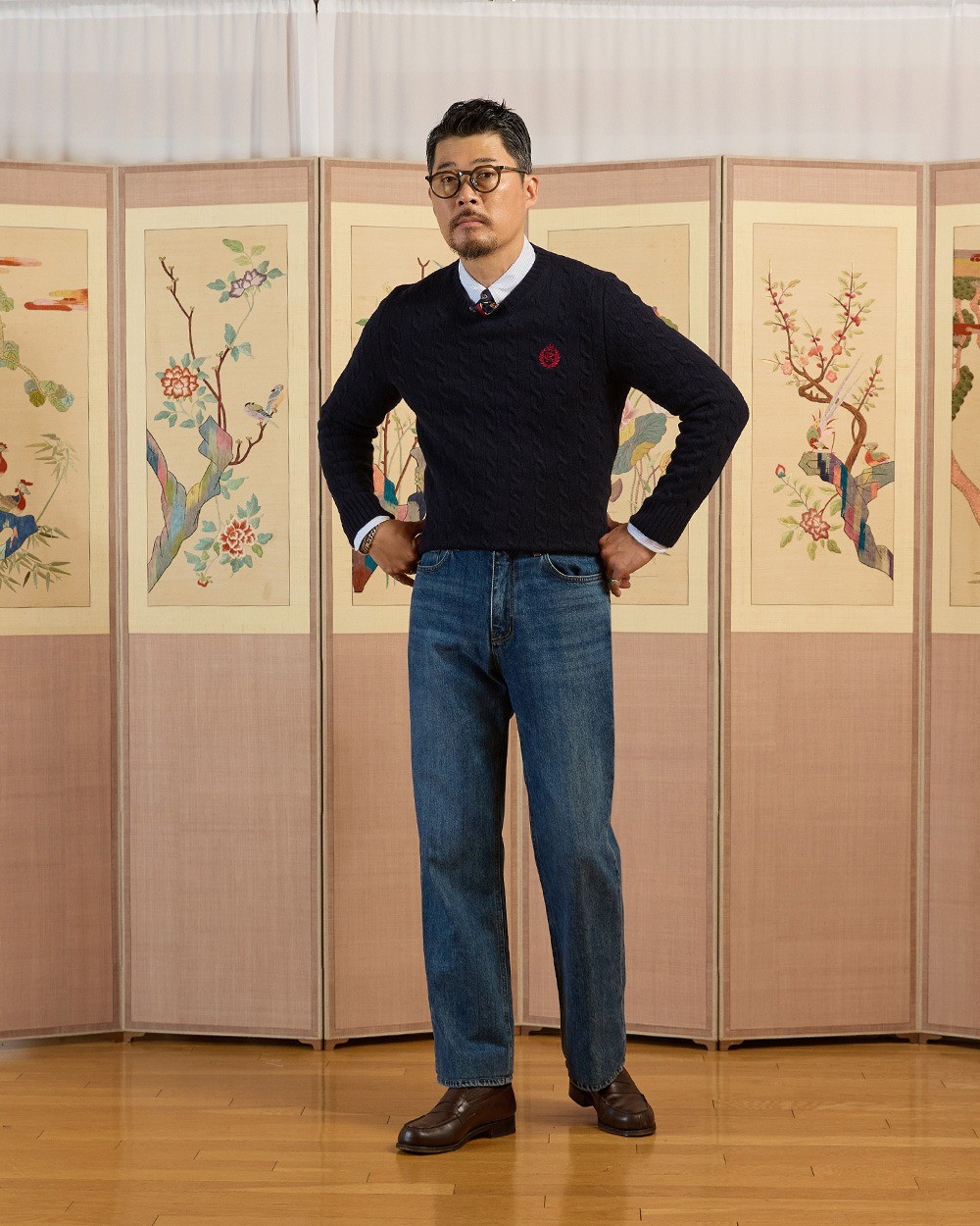 Man in a navy cable knit crewneck sweater over a white button-down collar, with mid-blue straight jeans and dark brown loafers, hands on hips in front of a folding screen