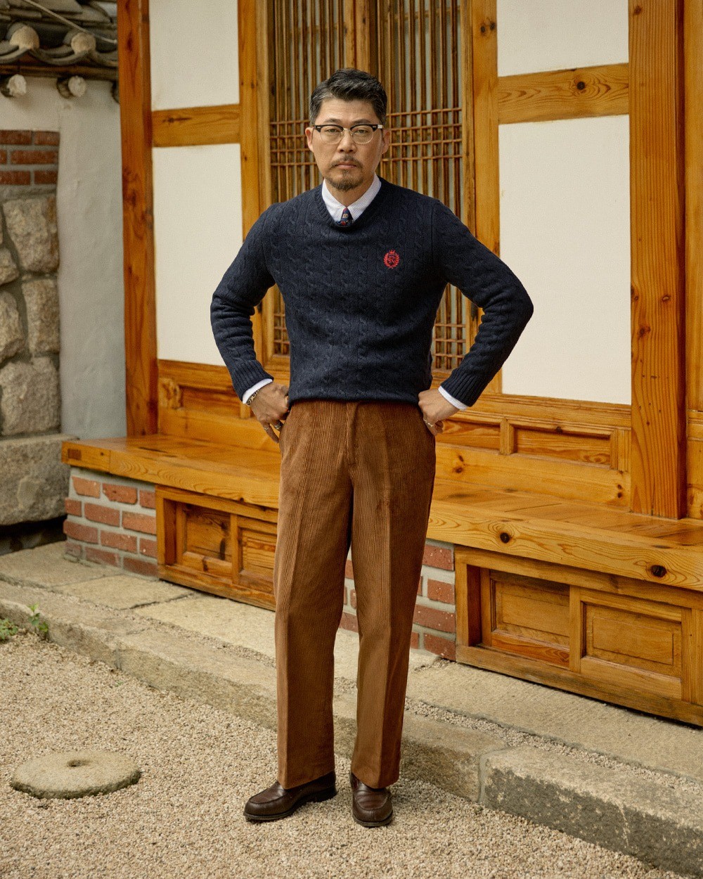 Man in a navy cable knit crewneck sweater over a button-down shirt and a tie, with wide brown corduroy trousers and dark brown loafers, standing in front of a hanok