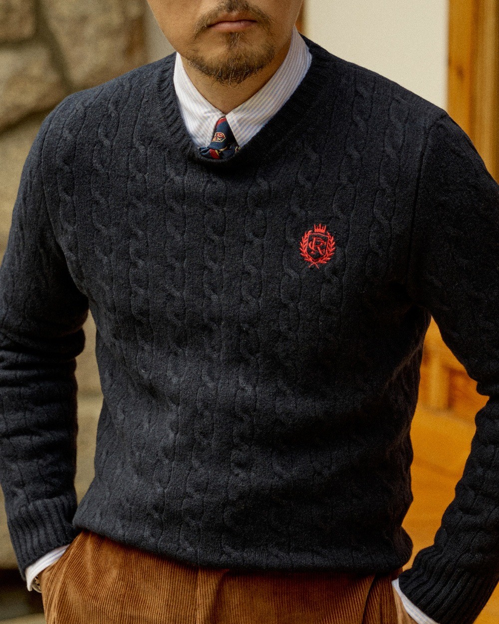Close-up of a navy cable knit crewneck sweater with a red embroidered crest, worn over a blue-and-white striped button-down collar and a patterned tie, above brown corduroy trousers