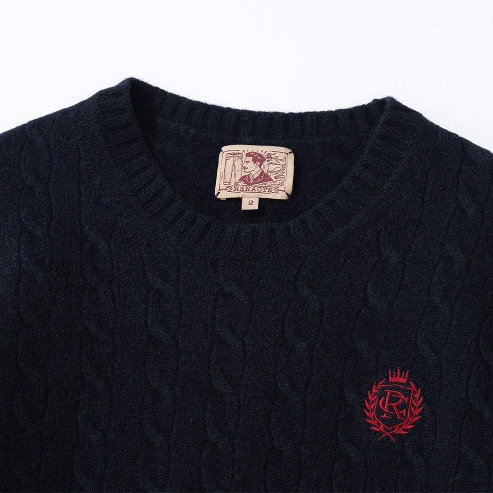 Neckline of a navy cable knit crewneck sweater laid flat, with a ribbed collar, a woven label inside the back of the neck and a small red embroidered crest on the chest