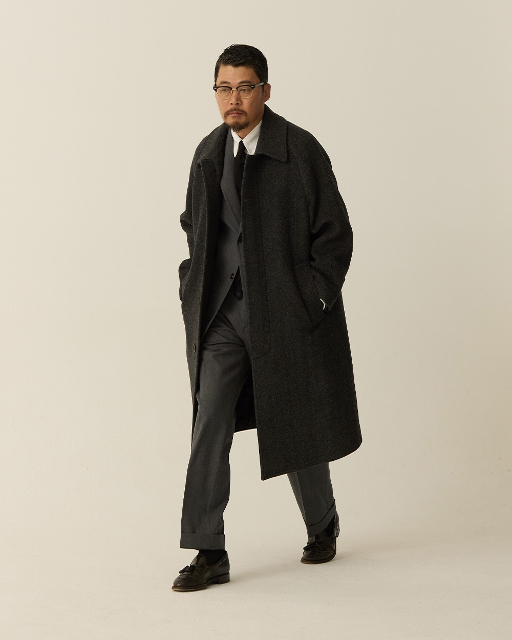 Man in a charcoal herringbone balmacaan coat worn open over a white shirt and tie with grey trousers and black shoes