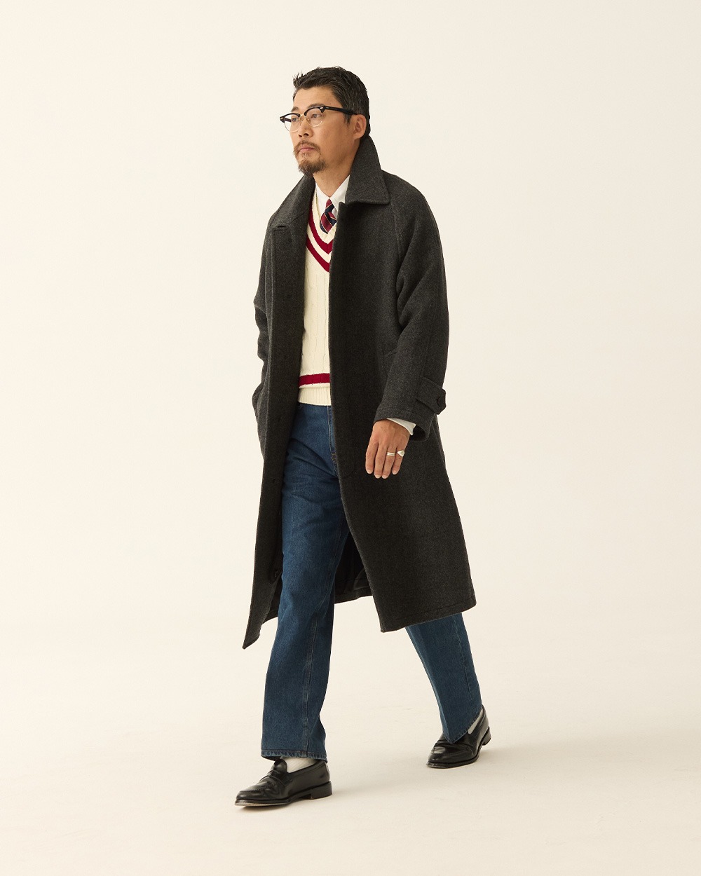 Man walking in a charcoal herringbone balmacaan coat worn open over a cream cricket sweater, with blue jeans and black loafers