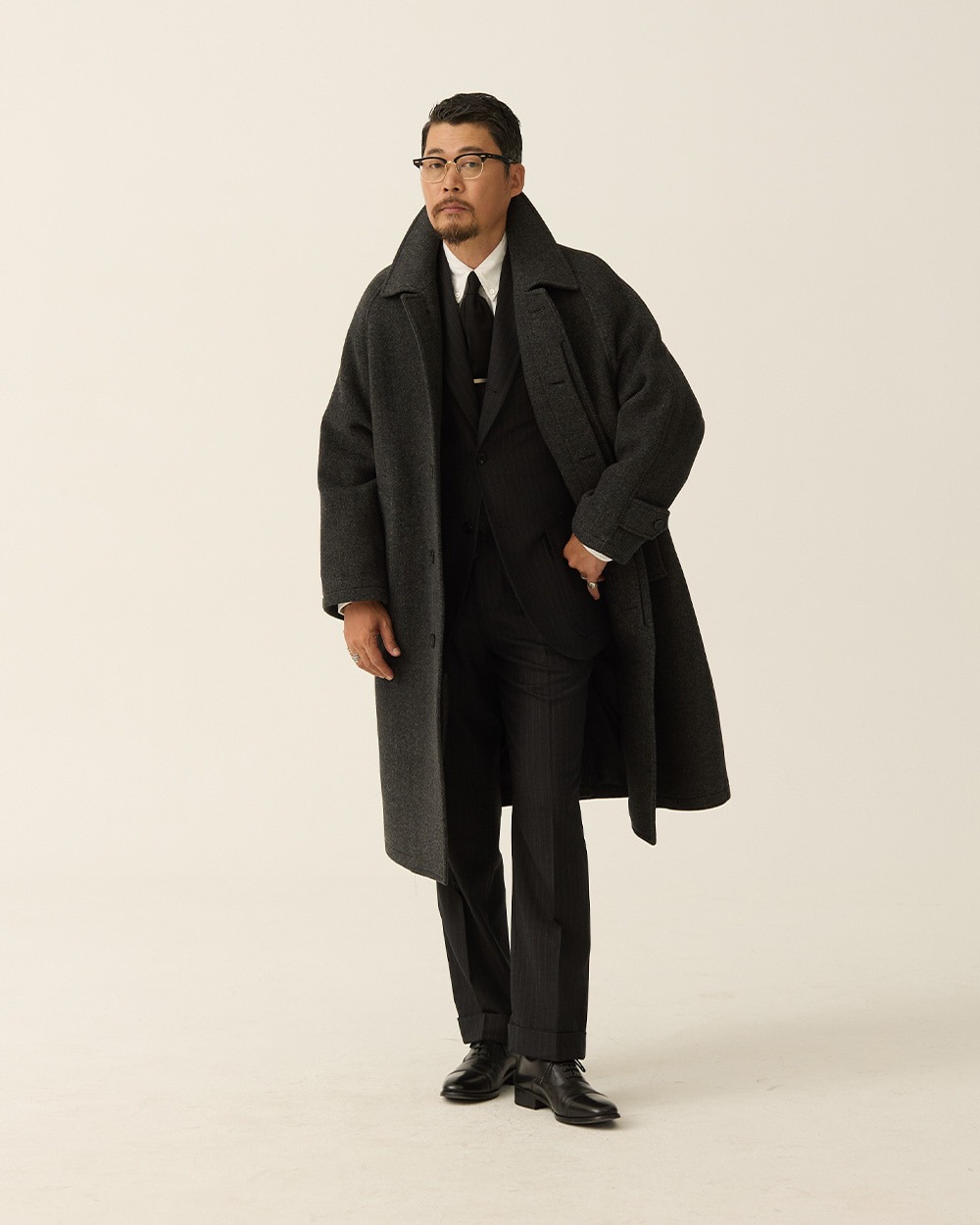 Man wearing a charcoal herringbone balmacaan coat open over a grey suit, white shirt and dark tie, hands in the coat pockets