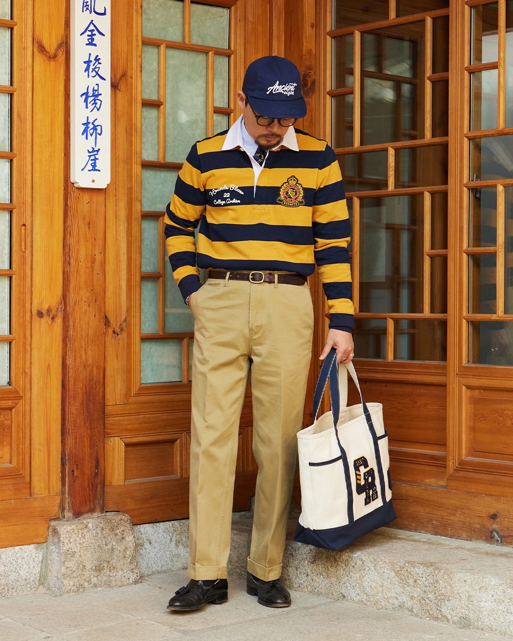 Man in a yellow and navy hooped rugby shirt with a navy baseball cap, khaki chinos, brown loafers and a canvas tote bag, standing in front of a hanok