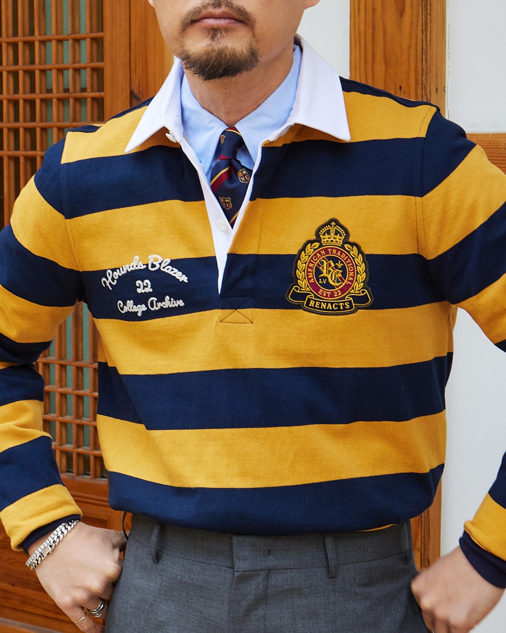 Close-up of a man in a yellow and navy hooped rugby shirt with an embroidered crest, worn over a light blue oxford shirt and a navy and yellow striped tie, with grey trousers