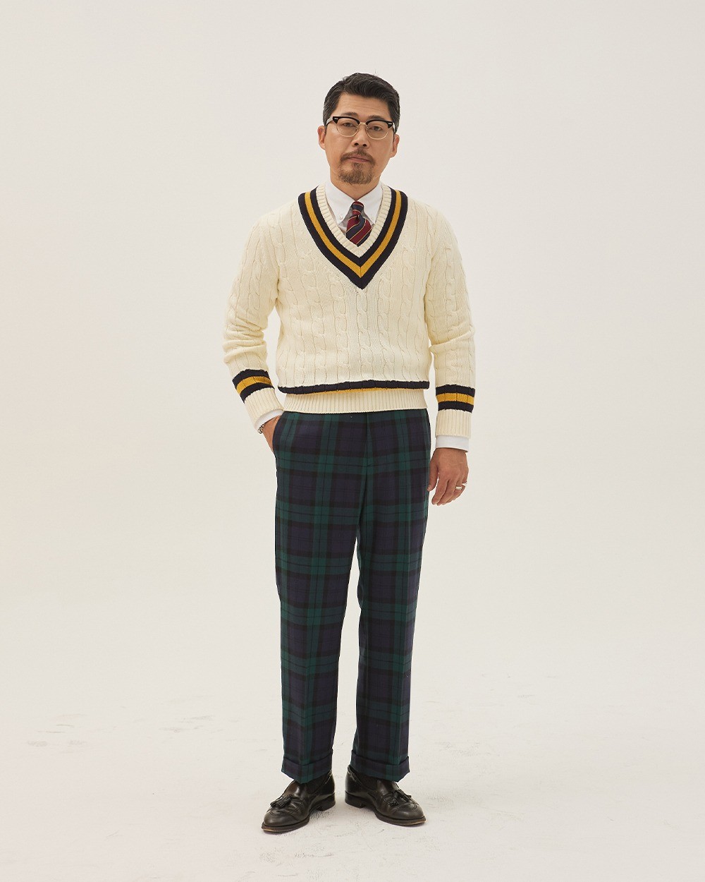 Man in an ivory cotton cricket sweater with navy and yellow stripes, a white button-down shirt and a red and navy striped tie, with Black Watch tartan trousers and black tassel loafers