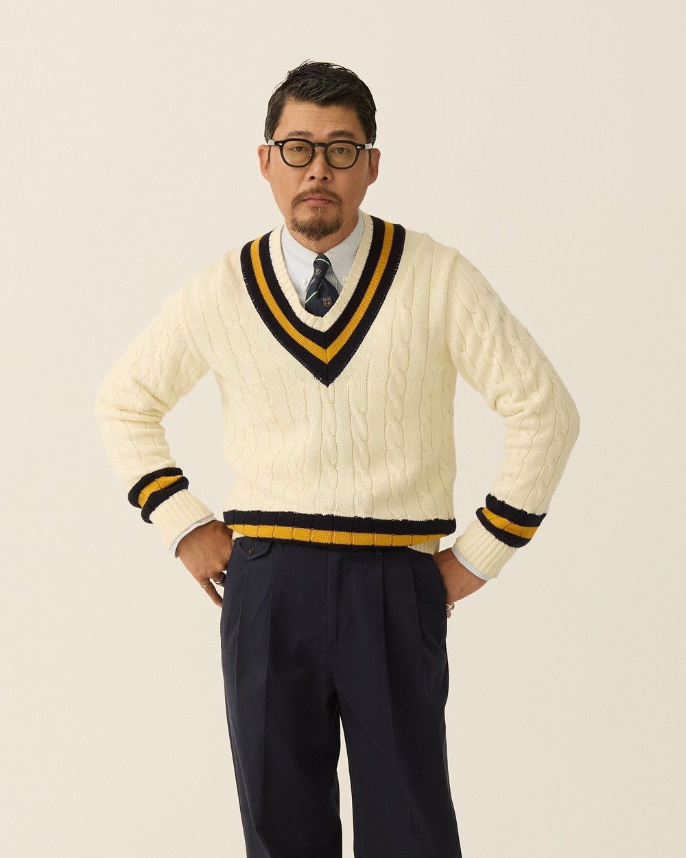 Man in an ivory cotton cricket sweater with navy and yellow stripes, a light blue button-down shirt and a navy patterned tie, with navy pleated trousers