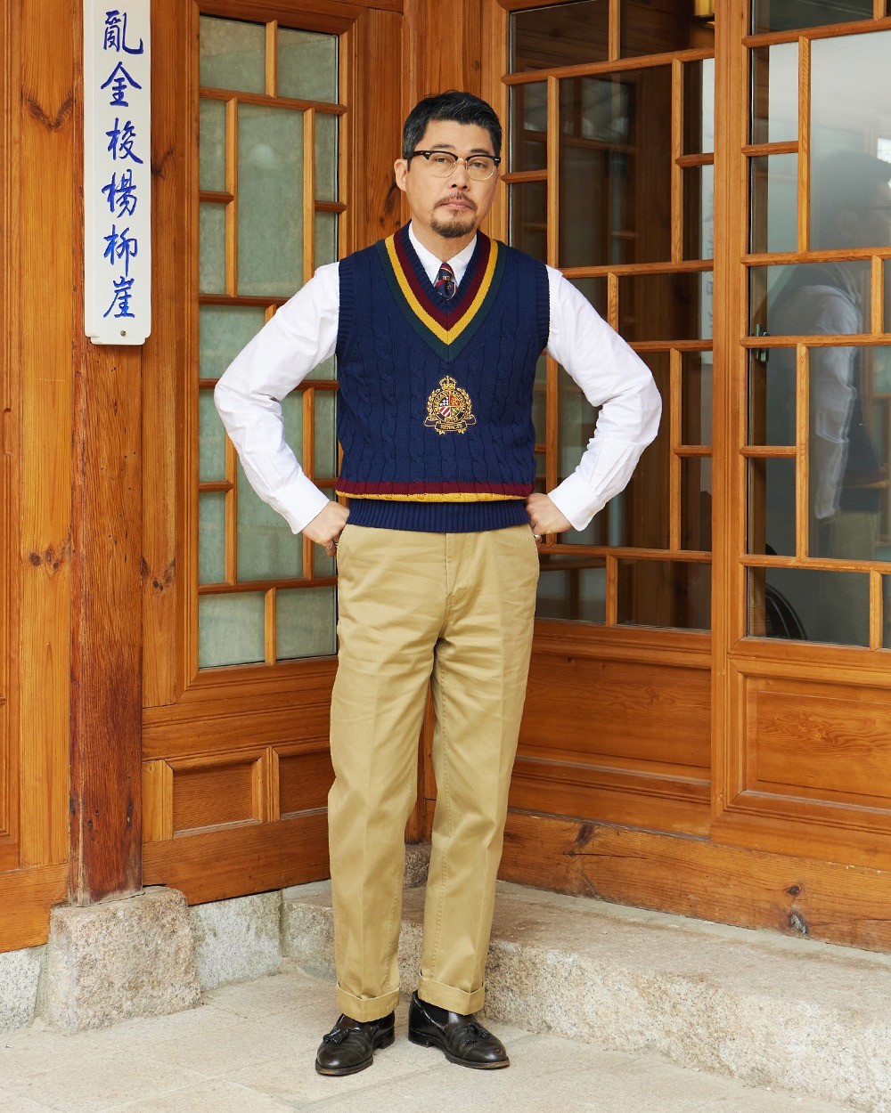 Man in a navy cotton cricket vest with a striped V-neck and an embroidered crest, over a white button-down shirt and a tie, with turned-up khaki chinos and black loafers