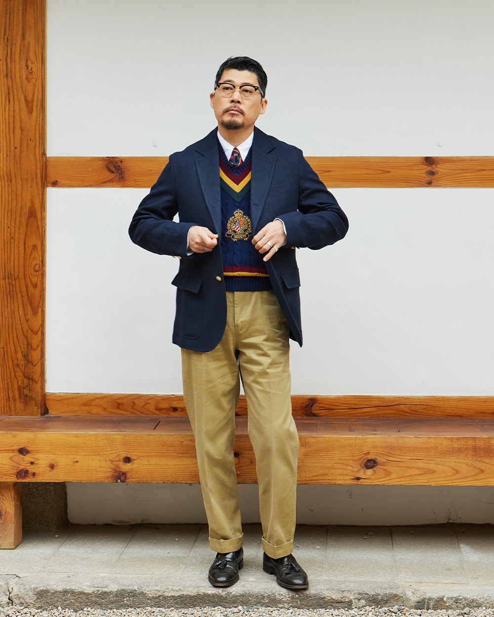 Man buttoning a navy blazer over a navy cotton cricket vest, a white button-down shirt and a tie, with khaki chinos and black loafers