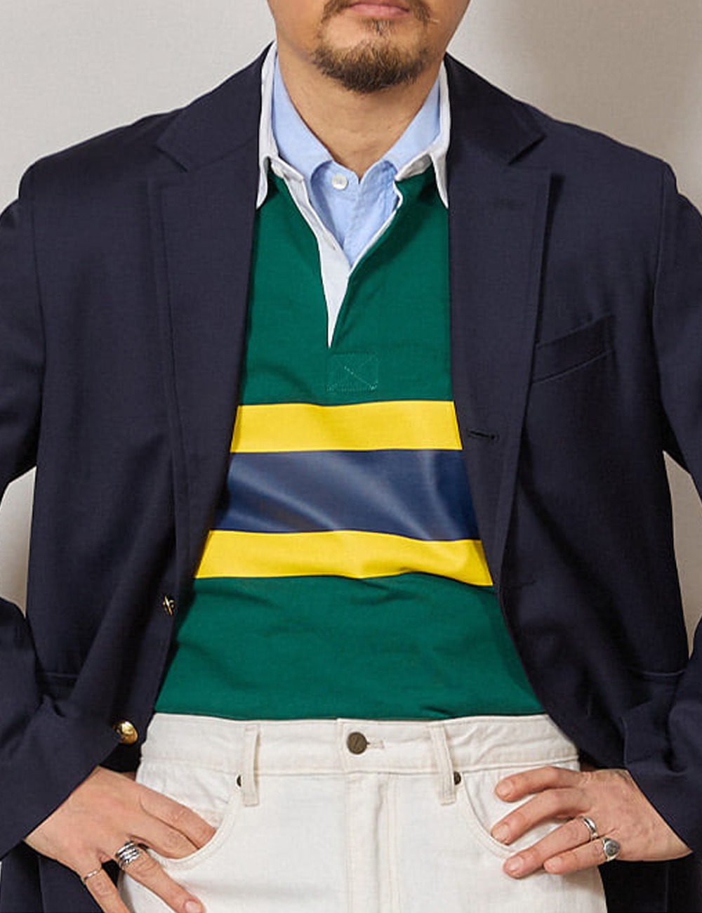Close-up of a navy blazer worn open over a green rugby shirt with a yellow and slate band, a light blue oxford shirt underneath, and white trousers