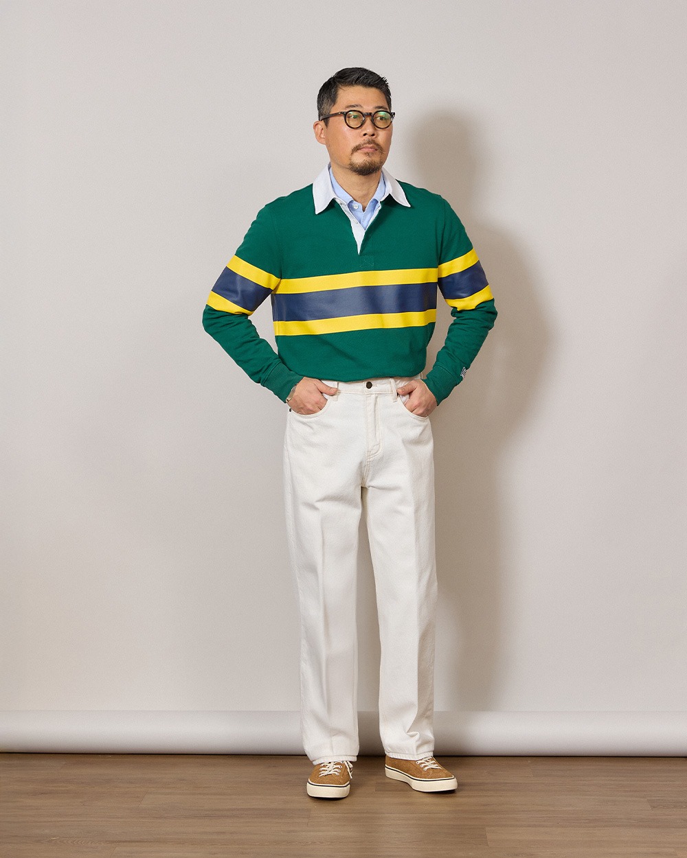 Man in a green rugby shirt with a yellow and slate band worn over a light blue oxford shirt, white jeans and cream canvas sneakers