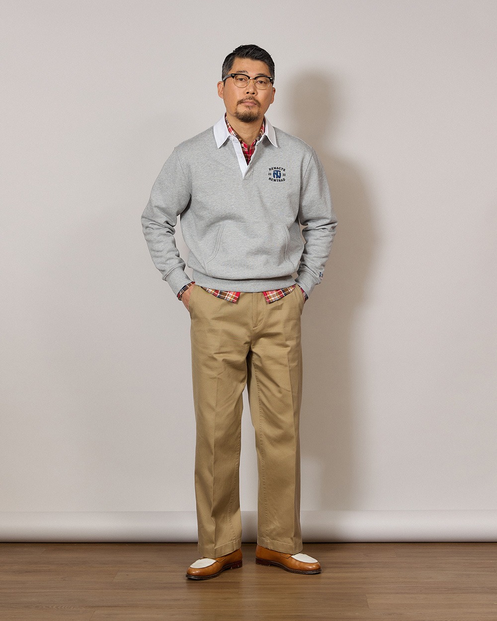 Man in a grey rugby collar sweatshirt worn over a red madras check shirt, with khaki chinos and brown loafers