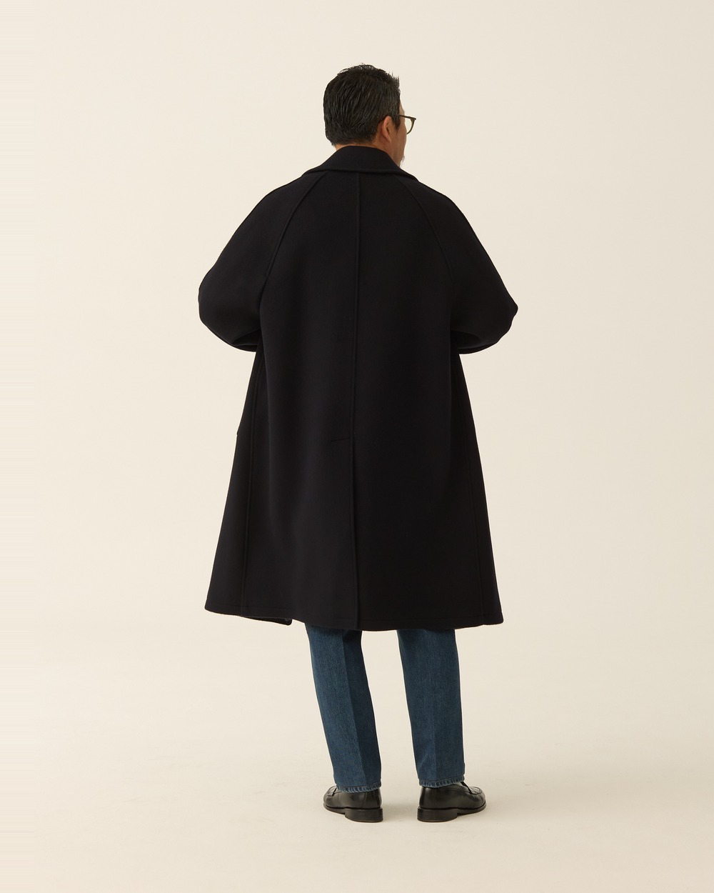 Back view of a man in a navy brushed wool balmacaan coat with jeans, showing the raglan shoulder seams and the single back vent