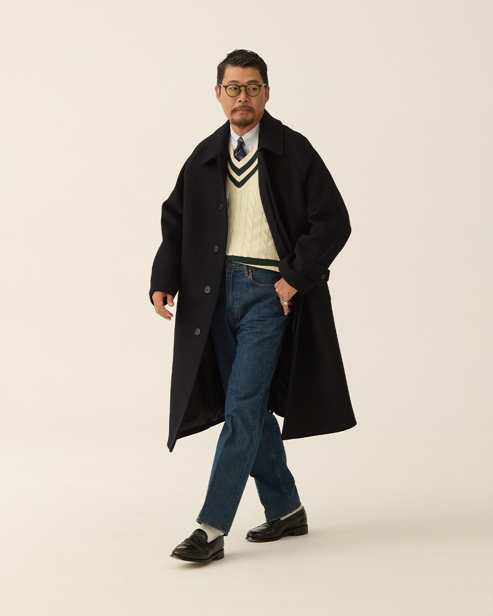 Man walking in a navy brushed wool balmacaan coat worn open over a cream cricket sweater and tie, with jeans and black loafers