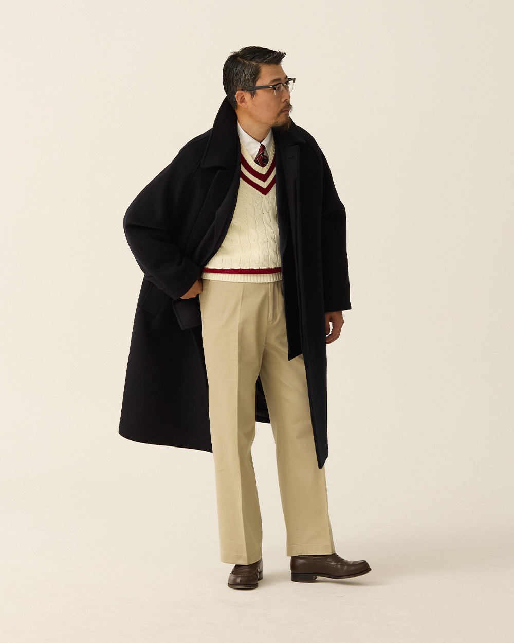 Man in a navy brushed wool balmacaan coat worn open over a cream cricket sweater, oxford shirt and tie, with khaki trousers and brown shoes