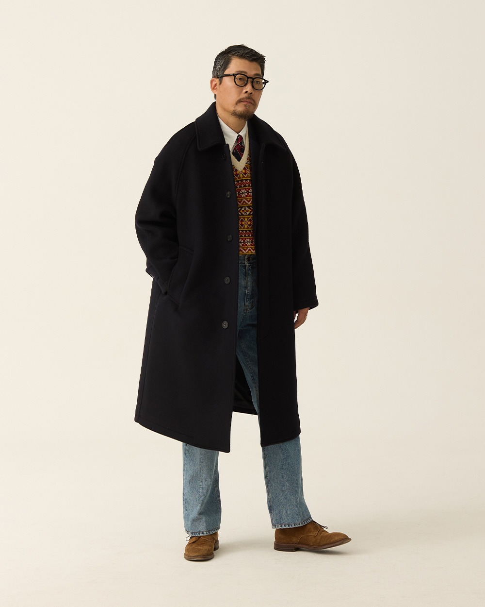 Man in a navy brushed wool balmacaan coat worn open over a Fair Isle vest and oxford shirt, with light-wash jeans and tan suede loafers