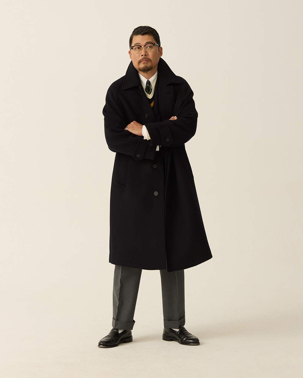 Man in a navy brushed wool balmacaan coat worn open over a grey suit, white shirt and striped tie, with black shoes