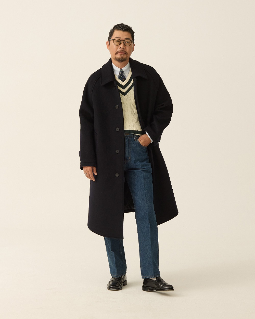 Man in a navy brushed wool balmacaan coat worn open over an oxford shirt and tie, with blue jeans and black loafers