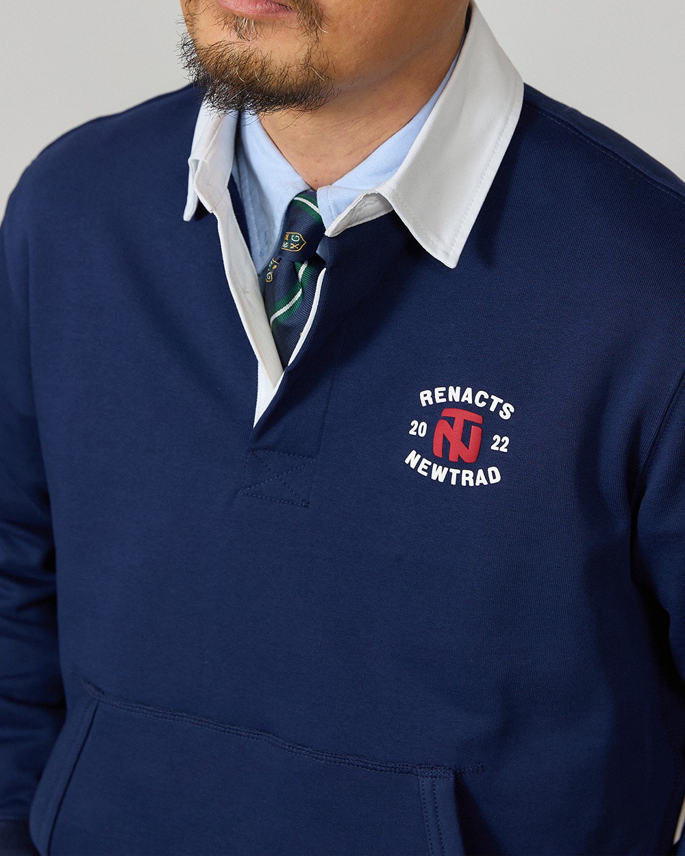 Close-up of a navy rugby collar sweatshirt's white collar over a light blue oxford shirt and a green and navy striped tie, with a small chest print