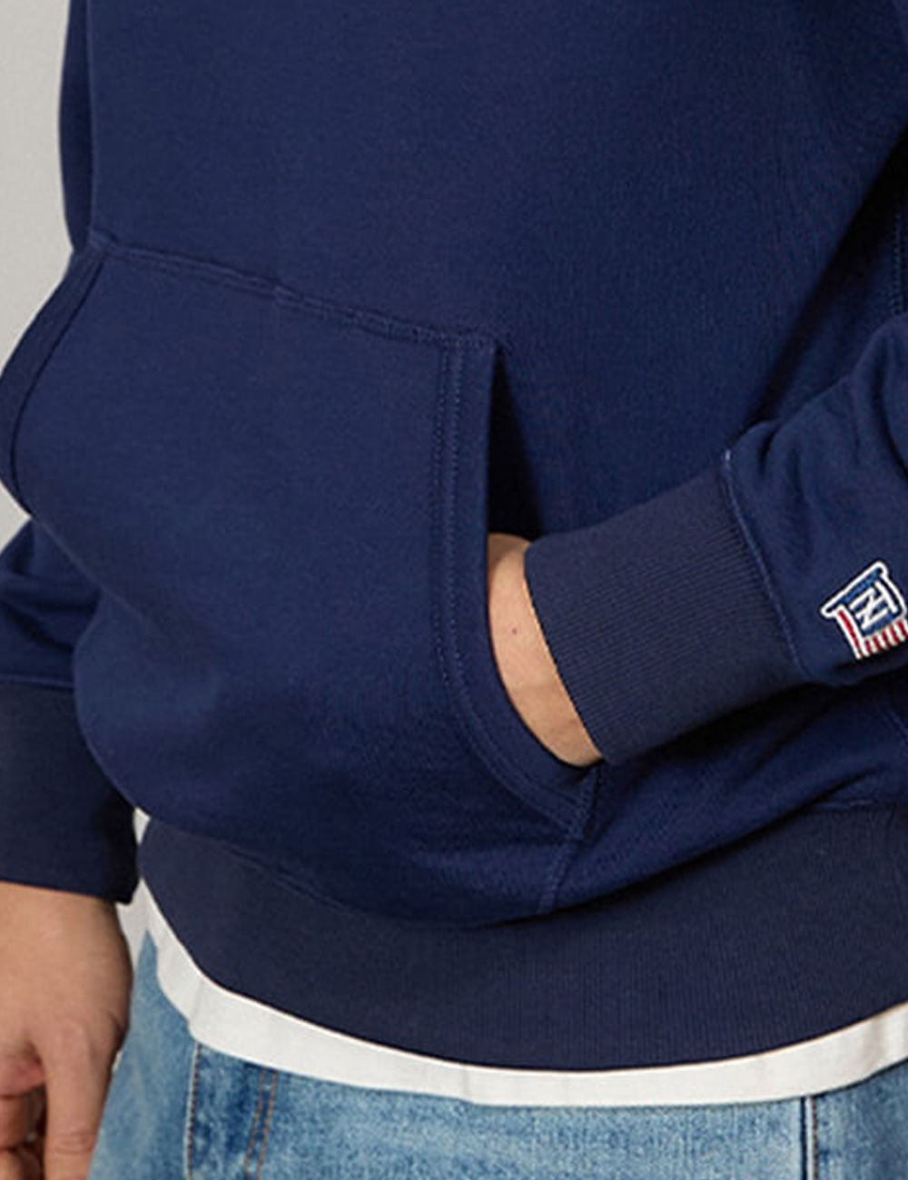 Close-up of a hand in the kangaroo pocket of a navy rugby collar sweatshirt, with a white T-shirt hem showing above light-wash jeans