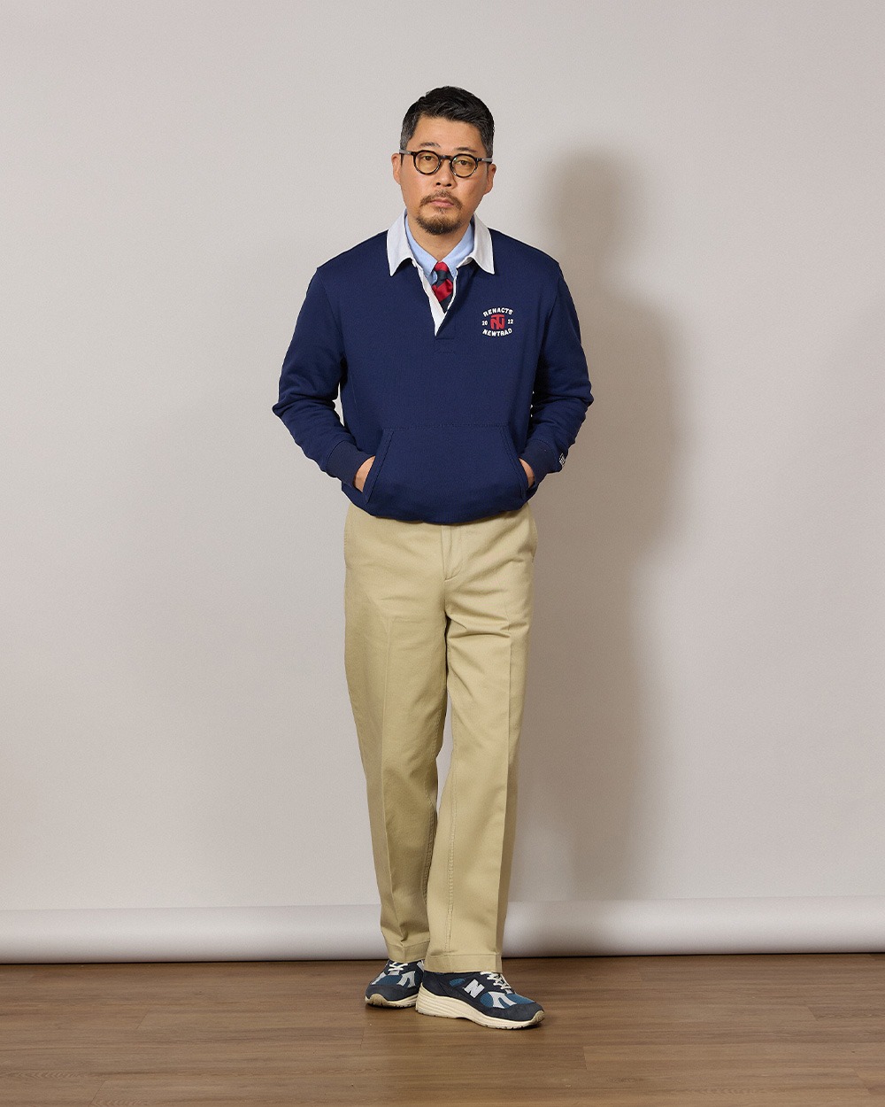 Man in a navy rugby collar sweatshirt worn over a light blue oxford shirt and a striped tie, with khaki chinos and navy canvas sneakers