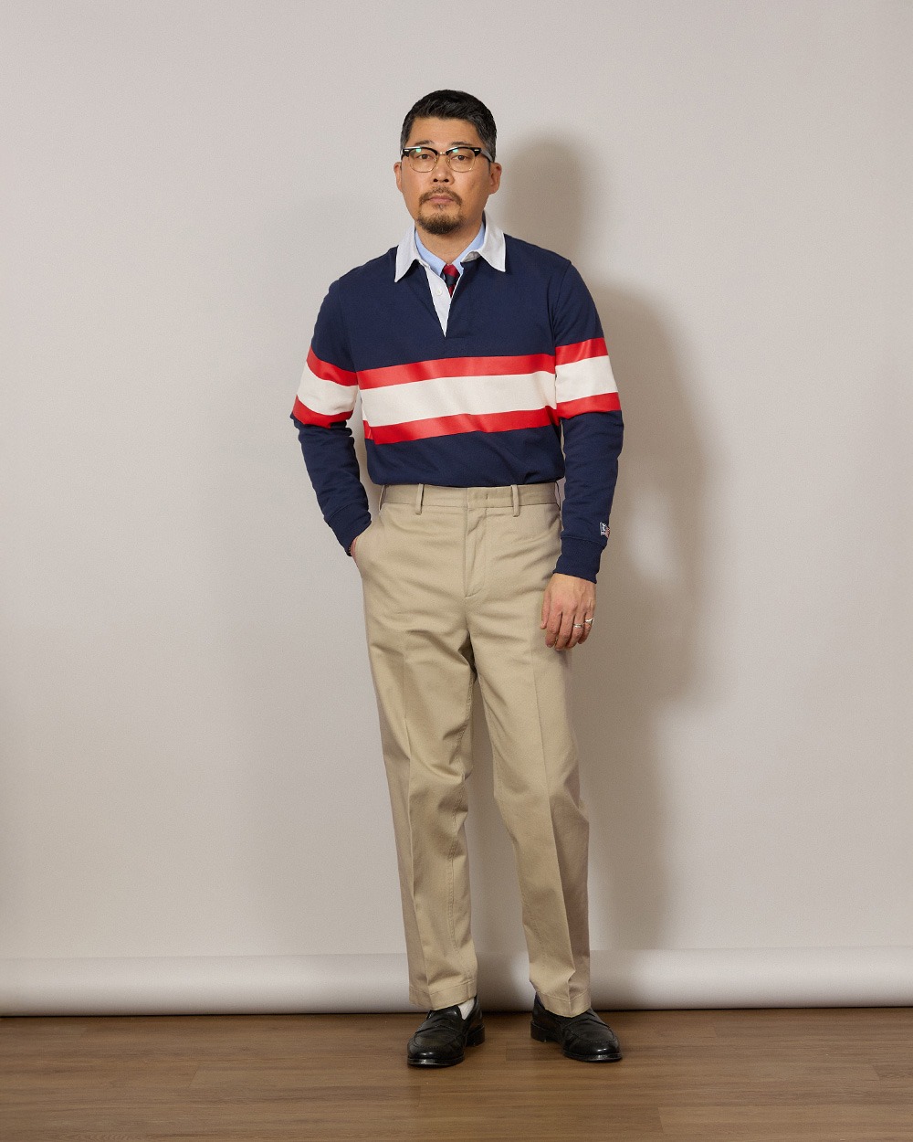Man in a navy rugby shirt with a red and white chest band, worn over a light blue oxford shirt and a tie, with khaki chinos and black loafers
