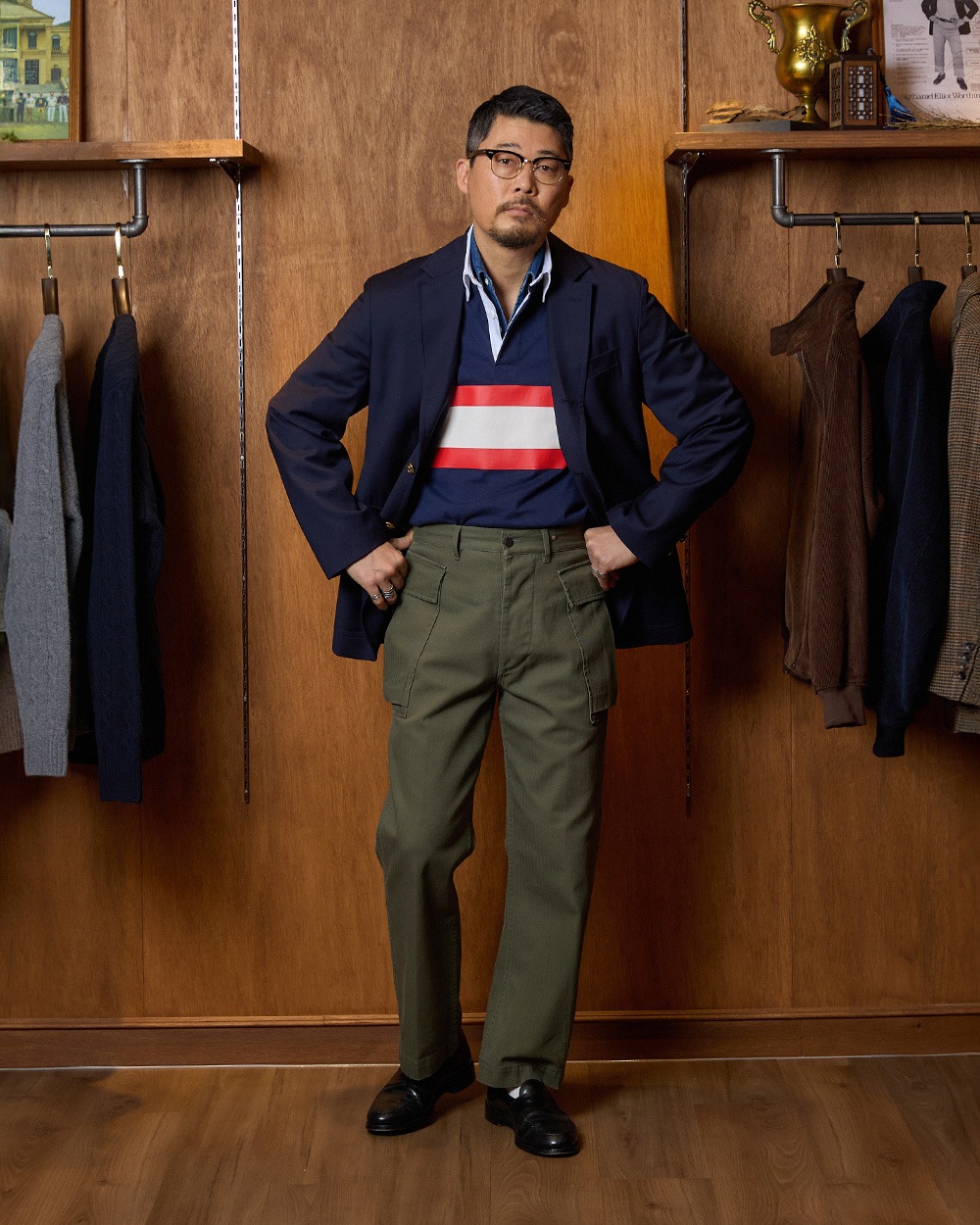 Man standing in a menswear shop wearing a navy blazer over a navy rugby shirt with a red and white chest band, olive trousers and black loafers