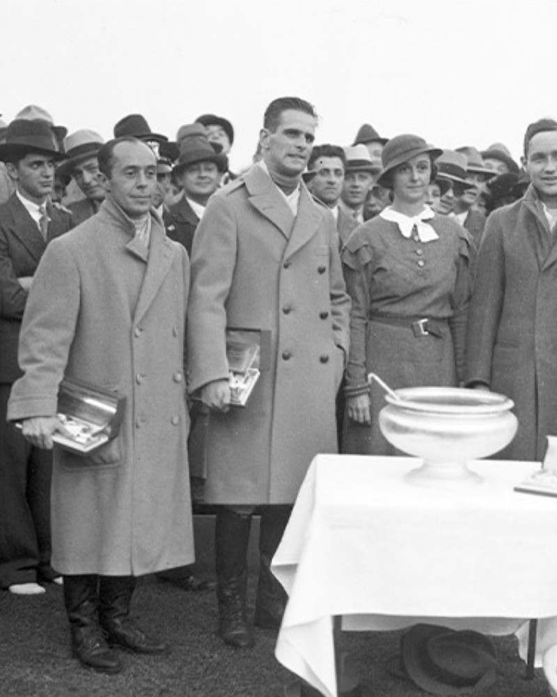 Black-and-white archive photograph of a crowd at an outdoor event, with a man in a light double-breasted overcoat in front and a silver trophy bowl on a white-clothed table