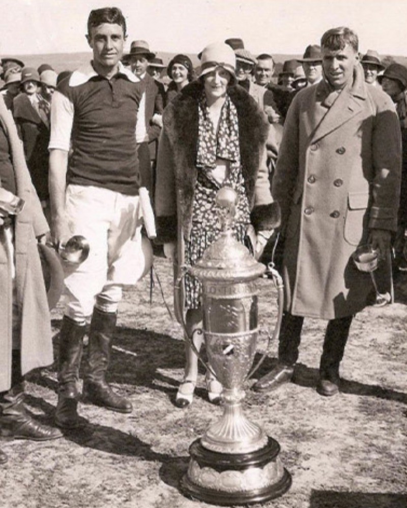 Black-and-white archive photograph of a polo player in a dark jersey and white breeches standing beside a large trophy cup, with a woman in a cloche hat and a man in a double-breasted overcoat behind him