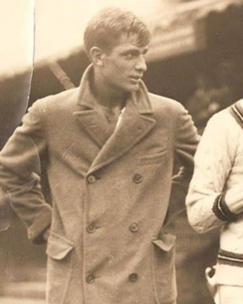 Sepia archive photograph of a young man in a double-breasted coat with wide lapels, standing outdoors with figures in white sweaters behind him