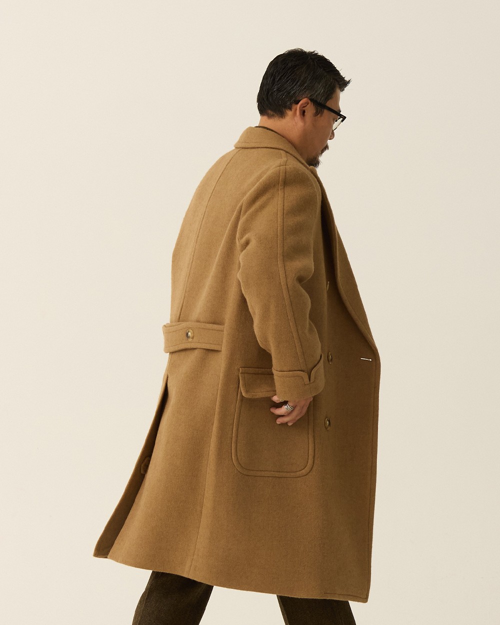 Back view of a man walking in a camel polo coat, showing the half belt, a patch pocket and the vent, with dark trousers