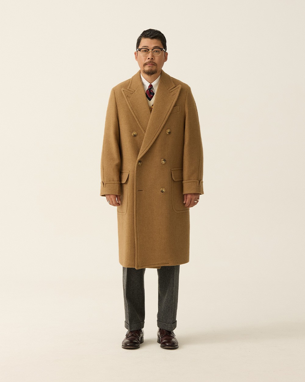 Man in a camel polo coat worn buttoned over a white shirt and striped tie, with grey flannel trousers and burgundy loafers, against a plain studio background