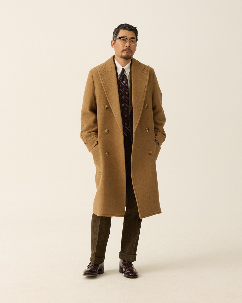 Man in a camel polo coat worn open over a dark suit, white shirt and patterned tie, with black loafers