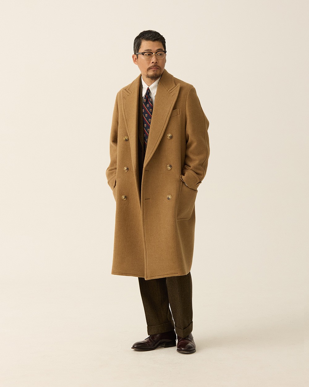 Man in a camel polo coat worn open over a dark suit and patterned tie, hands in the coat pockets, the coat's shoulder sitting outside the jacket's